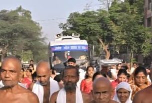 Crowd of people walking in a street procession, with some shirtless men in the foreground and a blue bus visible in the background.