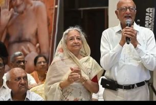 Elderly Indian couple on stage at an event; man speaks into a microphone while woman in a saree claps beside him.