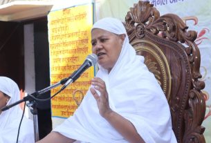 A nun or speaker in white robes speaks into a microphone while seated on an ornate wooden chair at a public event, with another woman in white nearby.