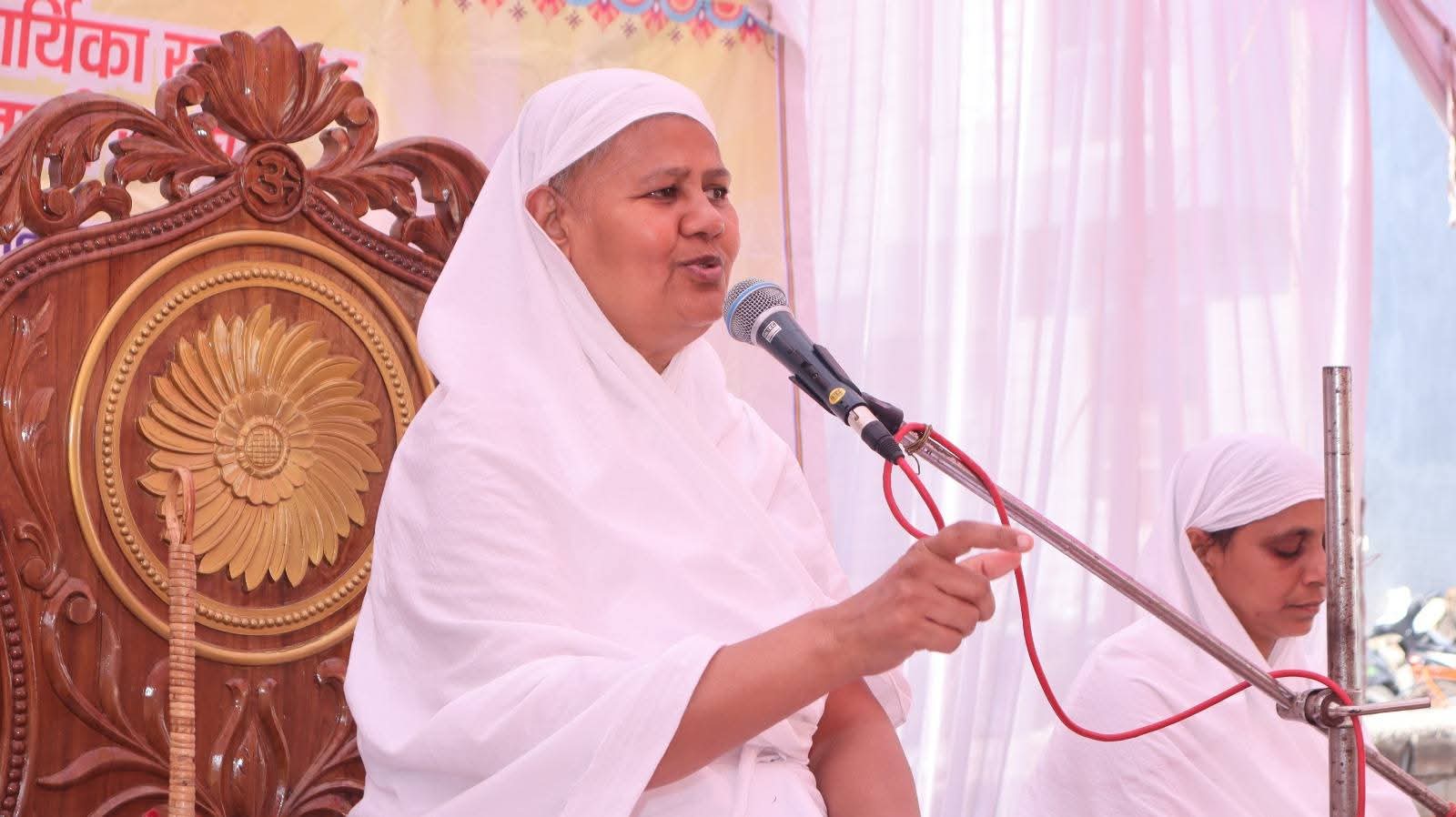 Woman in a white headscarf speaks into a microphone at a ceremony, seated on an ornate wooden chair; another woman sits nearby.
