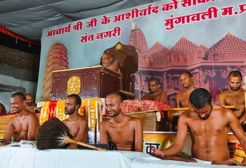 Group of shirtless men seated on stage during a Hindu religious ceremony, with a priest at a lectern and a temple backdrop behind them.