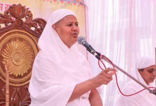 Woman in white robes speaks into a microphone on a stage, seated on an ornate carved wooden chair behind her.
