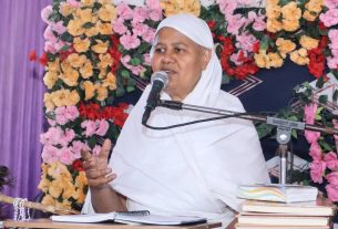Woman in a white hijab speaks into a microphone at a desk with books, against a colorful flower backdrop I
