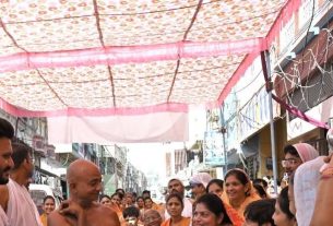 Crowded Indian street festival under a pink patterned canopy, with people in traditional attire gathered along the street.