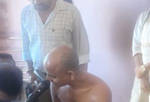 Group of adults in a clinic-like room watching a shirtless, bald man who speaks into a handheld microphone; medical equipment and cabinets visible in the background.