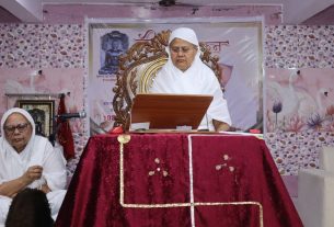 Senior nun in white habit stands at a wooden lectern delivering a talk, while another nun sits nearby in white robes.