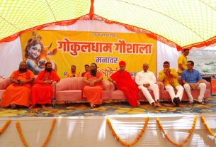 Panel of Hindu monks seated on pink sofas on a decorated stage under a bright tent; behind them is a large yellow banner with red Hindi script and images of a child and a cow.
