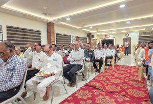 Group of men seated in rows at a conference hall, listening to a presentation; red patterned carpet runs down the center of the room.