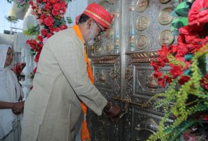 Man in traditional attire unlocks ornate silver temple doors amid red flower decorations, with a woman watching nearby.
