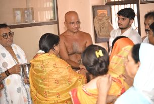 Group of people participating in a traditional Indian blessing ceremony; a shirtless man stands at center while others offer items and guidance.