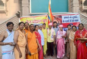 Group of Indian men and women in colorful traditional attire posing for a photo at a community event with banners in Hindi in the background.