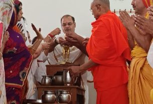 Group of people in traditional Indian attire gather around a small altar as a monk in orange robes blesses a ceremonial object while others offer prayers and hands are joined in reverence.
