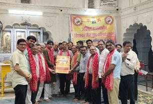Group of men in a ceremony, some wearing red scarves, posing with a framed certificate in a temple hall with a yellow banner in the background.