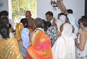 Crowd of people in traditional Indian attire gathered indoors for a ceremony, some with heads bowed and hands clasped.