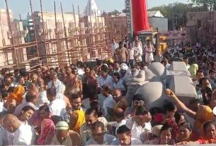 Large crowd in traditional clothing gathers around a vehicle in a street procession, with onlookers filling the area behind scaffolding and a tall red pillar in view.