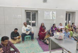 Group of men and women sit along a wall in a bright room, some reading papers, others waiting with colorful clothing on mats and chairs prep for a meeting/clinic.