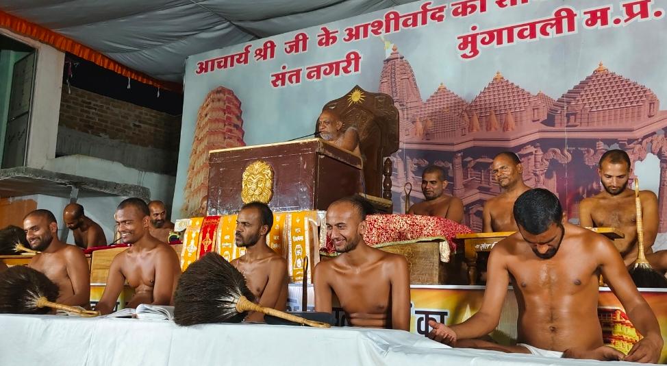 Monks participate in a temple ceremony; several shirtless men sweep with large brushes while a senior priest presides at a decorated podium, with a temple backdrop and Hindi banner.