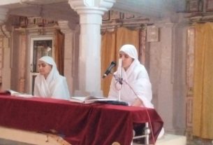 Two women in white head coverings sit at a red-draped table inside a temple, one reading from a book and the other speaking into a microphone.