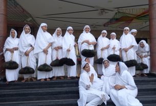 Group of women in white robes and head coverings standing on a staircase, some holding wooden staffs, indoors for a ceremony.",