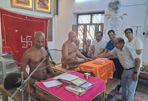 Monks conducting a Hindu puja ceremony in a small temple room with assistants handling ritual items and onlookers nearby.