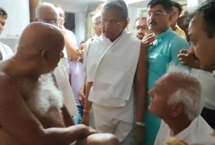 Group of men in white robes surround a shirtless elderly man with a long white beard in what appears to be a religious or ceremonial gathering indoors.
