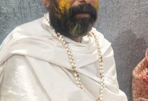 Man in white robes with a rainbow turban, red tilak on the forehead, yellow powder on the cheeks, wearing a beaded necklace.