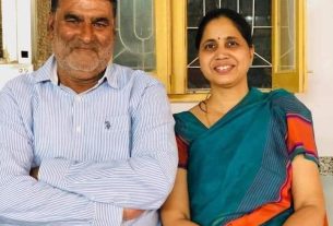 Smiling middle-aged man in a striped shirt and a woman in a teal sari sit together in front of a window.