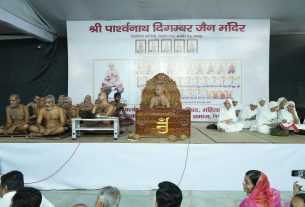 Monks seated on stage for a ceremony, with a large ornate throne and a Hindi banner behind them; audience seated in front.