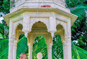Nude man meditating at a white marble shrine with a domed roof, surrounded by palm trees and string lights.