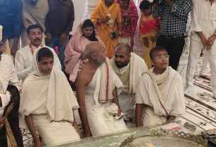 Group of people in traditional Indian attire gathered for a wedding ceremony, seated on the floor around a ritual setup and a seated elderly man with a white dhoti and red border.