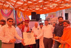 Group of men in traditional and formal attire at a ceremonial event, presenting a wrapped plaque on a stage under an orange fabric canopy.