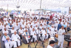 Large outdoor gathering of men in white attire seated under a white tent, with a speaker and cameras at the front.