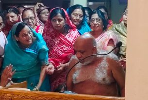 Shirtless, bald man with white ash on his chest sits at a carved wooden podium with a microphone, surrounded by women in colorful saris at a ceremony.
