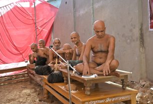 Group of shirtless men sitting cross-legged on wooden platforms at an outdoor cremation site, with red fabric tent in the background.