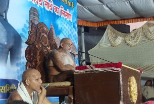 Bald monk seated at a carved wooden throne-like chair on stage, podium with red cloth, blue banner backdrop with Hindi text, in a tented event space.