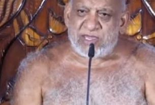 Elderly, bald man with a gray beard speaks into a microphone, shirtless, against an ornate wooden backdrop.