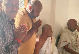 Group of men seated indoors, several with hands in prayer and bowed heads, wearing white robes and casual clothing.