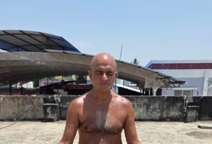 Bald shirtless man meditating cross-legged on a sunlit rooftop with concrete walls and a large curved canopy behind him, blue sky above