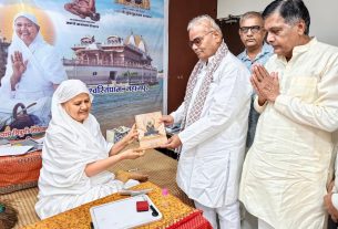 An elderly woman in white hands a diary to an elderly man while two men stand nearby, in front of a large poster with a Hindu temple image.