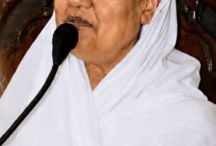A woman in white traditional attire speaks into a handheld microphone at a public event.