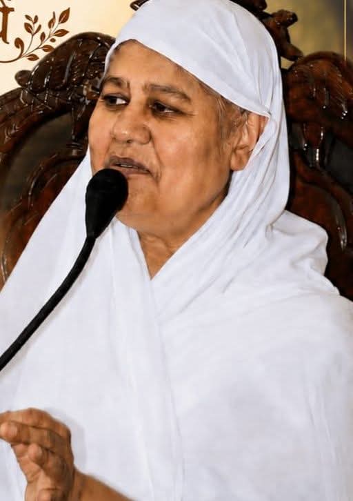 A woman in white traditional attire speaks into a handheld microphone at a public event.
