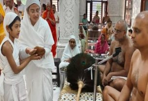 Young boy in white offers a coconut to a priest during a Hindu temple ceremony, with monks seated nearby and other worshippers in the background.
