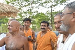 Group of men outdoors, some wearing orange robes, smiling and chatting near a thatched shelter and trees in the background.