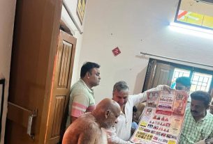 Shirtless elderly man seated with a group, looking at a large informational poster held by another man in a white shirt.",