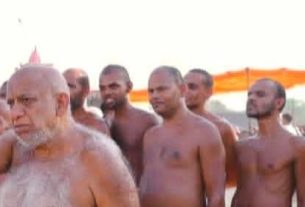 Elderly bearded sadhu raises his hand among a group of shirtless men at an outdoor religious gathering.