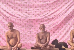 Five bare-chested men sit cross-legged on a wooden platform, meditating in front of a pink patterned backdrop.