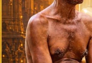 Shirtless elderly man with a lean, weathered frame sits in profile against a gold-toned temple interior, suggesting a ceremonial or religious setting.