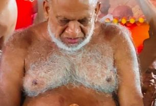 Elder man with white beard, shirtless, eyes down, holding a wooden cane in a bright festival setting.