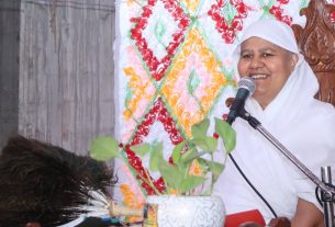 Woman in a white headscarf speaks into a microphone while seated on a carved wooden chair, with a colorful quilt backdrop.