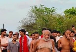 A crowd of men walking outdoors, several shirtless, with an older bearded man at the center; trees and blue sky in the background.
